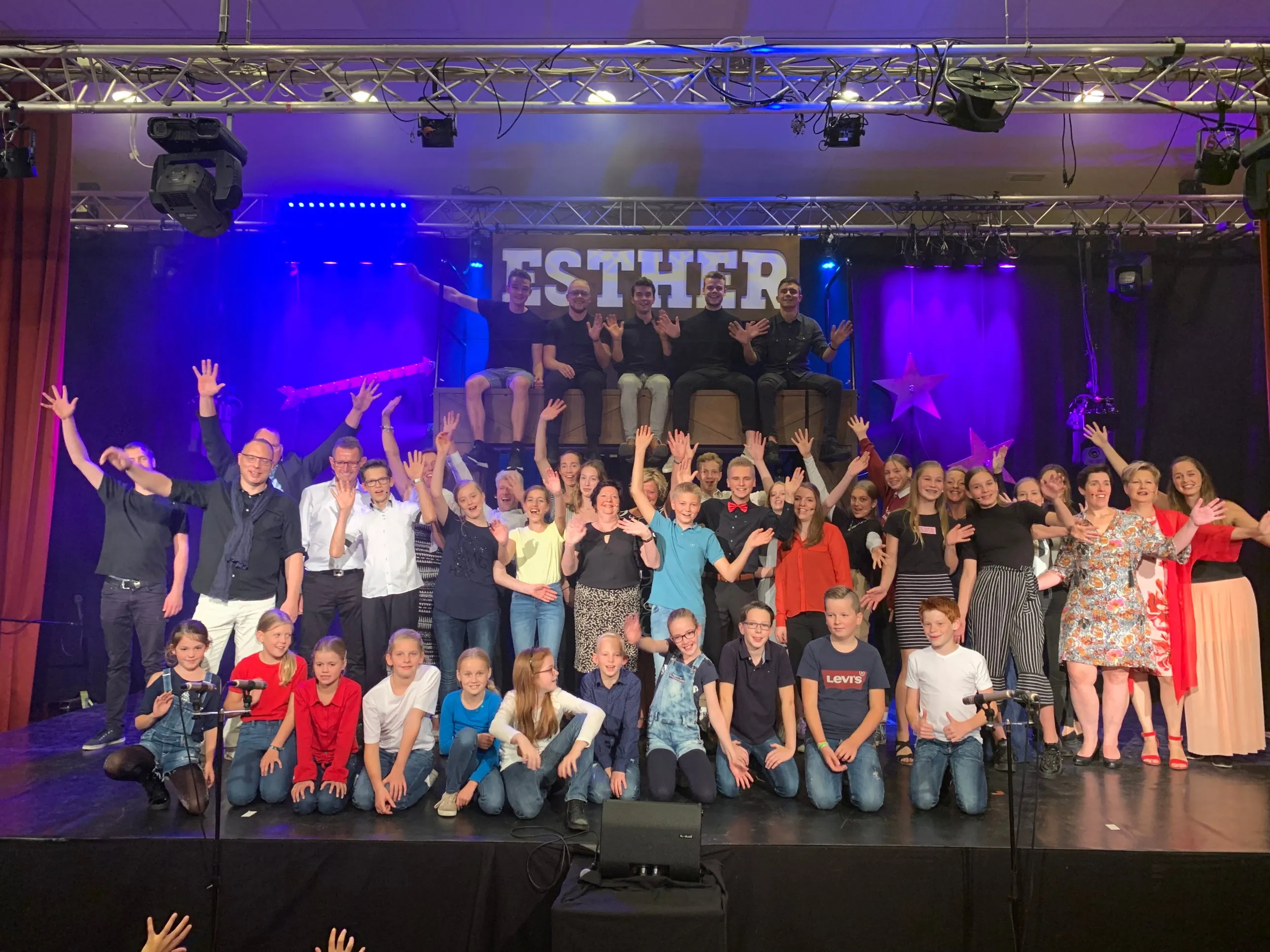 Het hele ensemble van de musical Esther zwaait vanaf het podium, van kinderen tot volwassenen, onder paars en blauw podiumlicht
